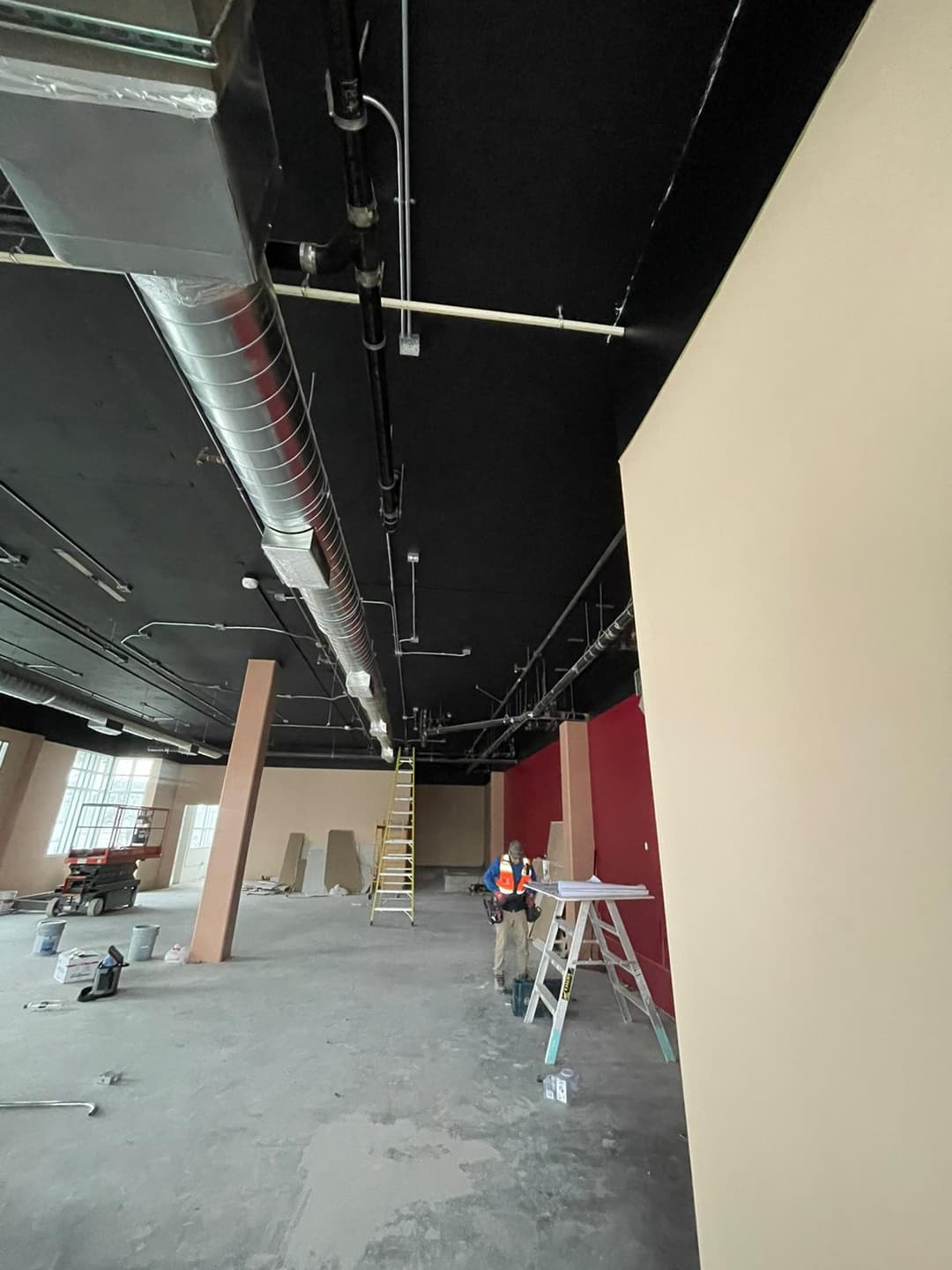 Construction site showing a worker on a ladder with exposed ducts and unfinished walls.